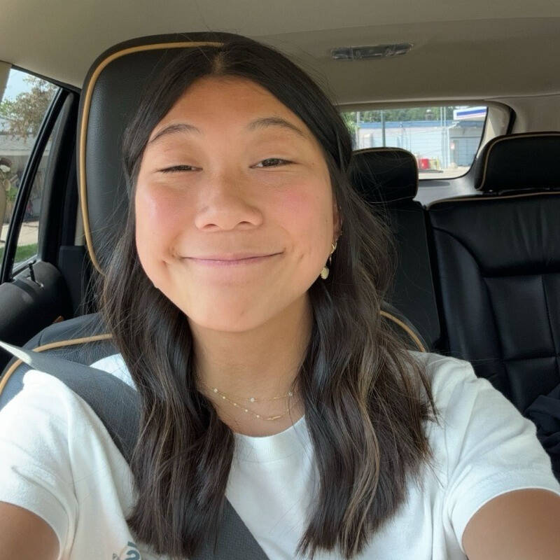 Clara Brooks, is sitting in the front seat of a car, wearing a white t-shirt and a delicate layered necklace. The car interior features black seats with tan piping, and there is a seatbelt across her chest.